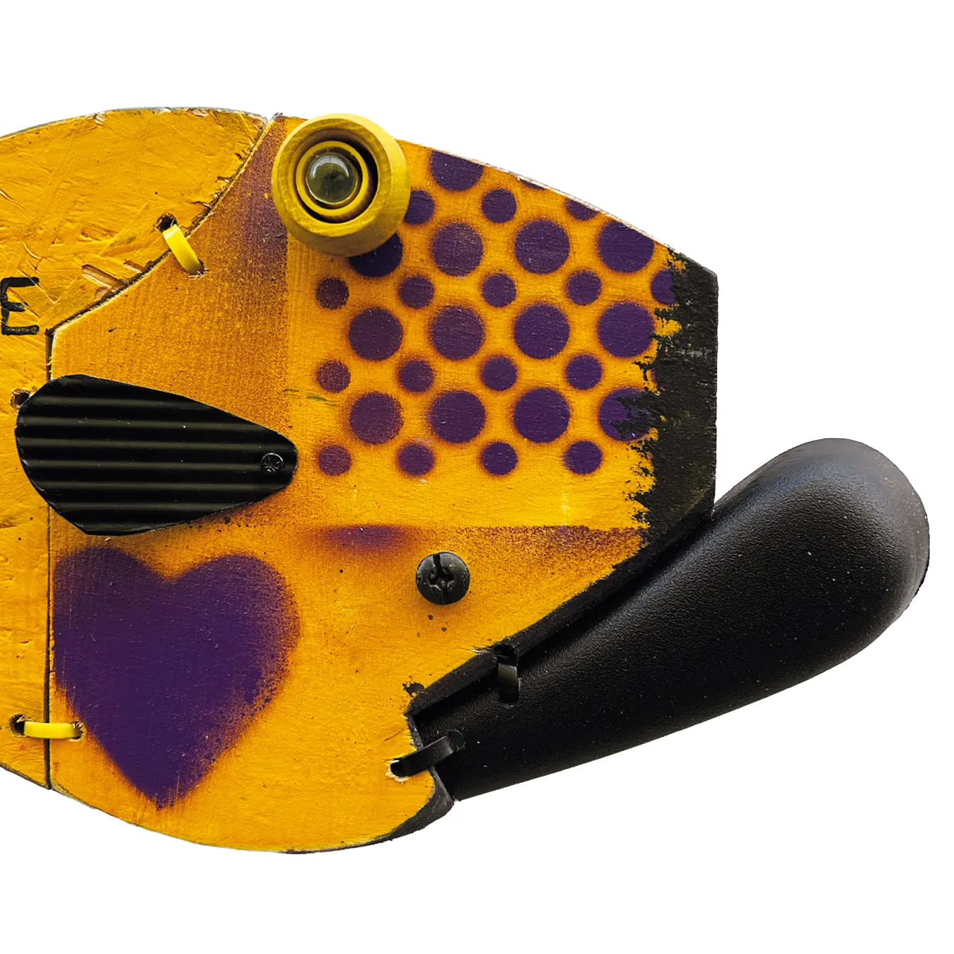 Dettaglio della testa del pesce in legno giallo con occhio sporgente, cuore dipinto in viola, motivi geometrici a pallini e manico nero come bocca