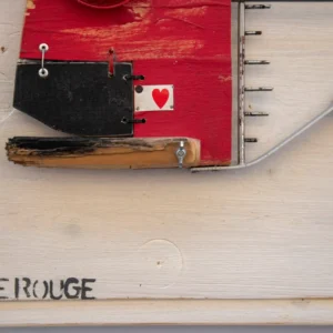 Dettaglio della scultura su pannello di legno chiaro intitolata 'Tête Rouge' del 'P.F. 2025'. Presenta una grande testa in legno rosso acceso con un occhio ricavato da un vistoso tappo di plastica rosso a raggiera. La mascella inferiore è un blocco nero con un piccolo riquadro bianco contenente un cuore rosso. Il corpo è una struttura filiforme di metallo che racchiude una fila di chiodi-lisca e termina con una coda a mezzaluna nera.