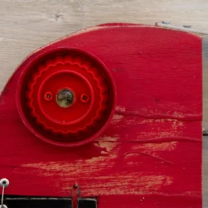 Dettaglio della scultura su pannello di legno chiaro intitolata 'Tête Rouge' del 'P.F. 2025'. Presenta una grande testa in legno rosso acceso con un occhio ricavato da un vistoso tappo di plastica rosso a raggiera. La mascella inferiore è un blocco nero con un piccolo riquadro bianco contenente un cuore rosso. Il corpo è una struttura filiforme di metallo che racchiude una fila di chiodi-lisca e termina con una coda a mezzaluna nera.