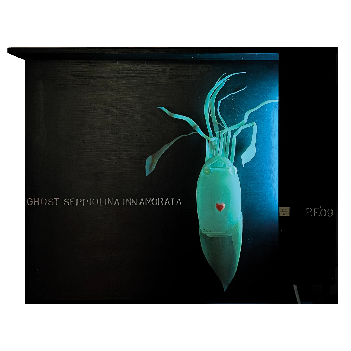 Una scultura retroilluminata di una seppia stilizzata, realizzata con materiali plastici traslucidi, è montata su un pannello di legno scuro all'interno di una cornice a scatola nera. Il corpo traslucido della seppia si illumina di un bagliore verde-azzurro al neon, rivelando i tentacoli arricciati, due occhi sferici e un piccolo cuore rosso al centro del corpo. A sinistra, la scritta bianca "GHOST SEPPIOLINA INNAMORATA"; a destra, la firma dell'artista "P.F. '09" e un piccolo simbolo della lampadina. Lo sfondo scuro esalta il contrasto luminoso