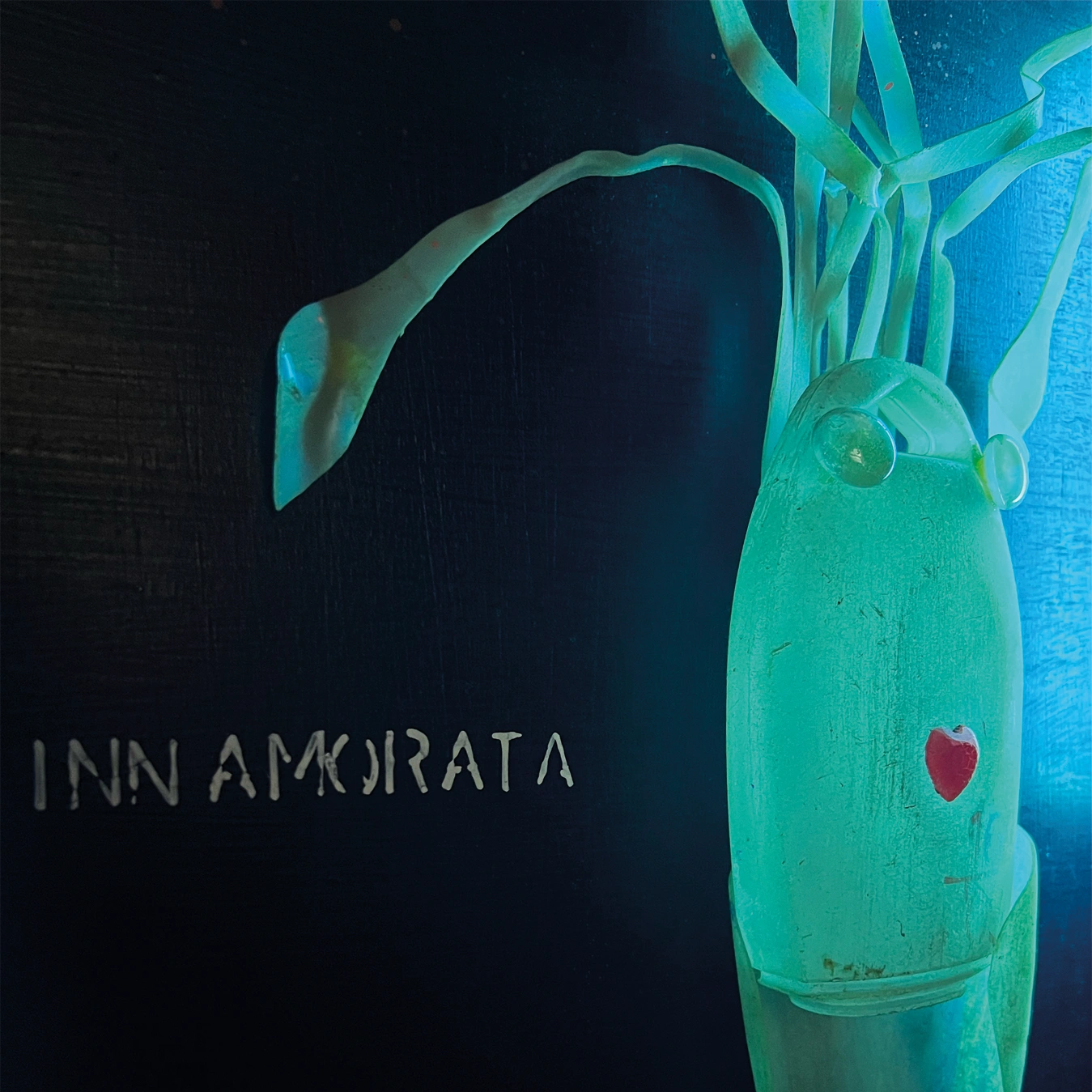 Una vista laterale ravvicinata della seppia stilizzata retroilluminata di verde-azzurro. Il corpo traslucido brilla intensamente. Si notano il piccolo cuore rosso e gli occhi sferici. A sinistra, parte della scritta bianca sul legno scuro: "...INNAMORATA". La luce al neon illumina i contorni dei tentacoli.