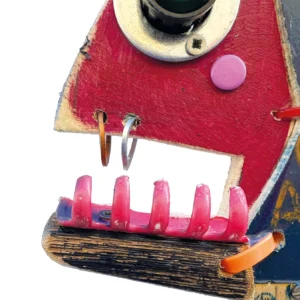 Dettaglio della bocca di una scultura di pesce in legno con denti fatti da molletta fucsia e anelli metallici come piercing sul labbro superiore