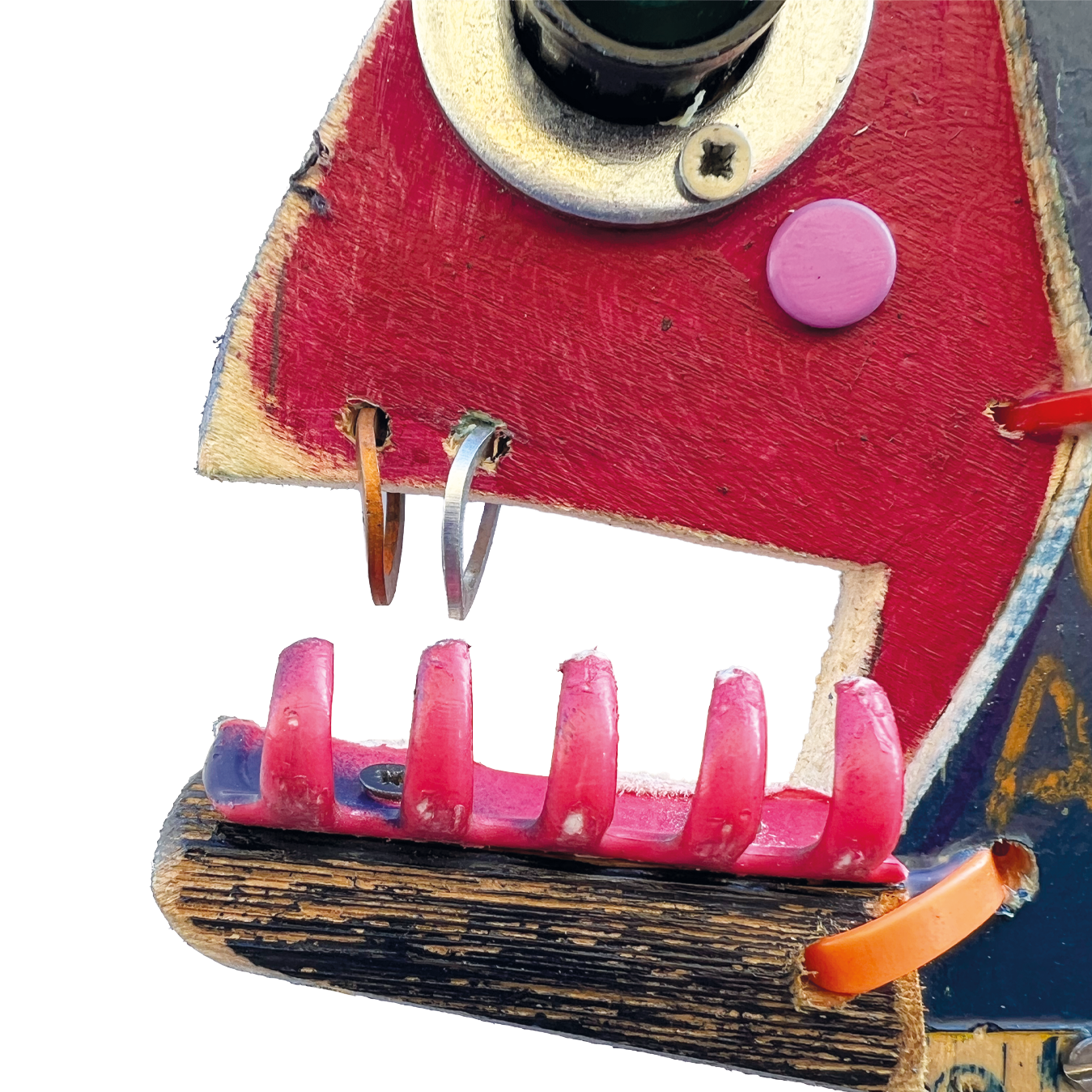 Dettaglio della bocca di una scultura di pesce in legno con denti fatti da molletta fucsia e anelli metallici come piercing sul labbro superiore